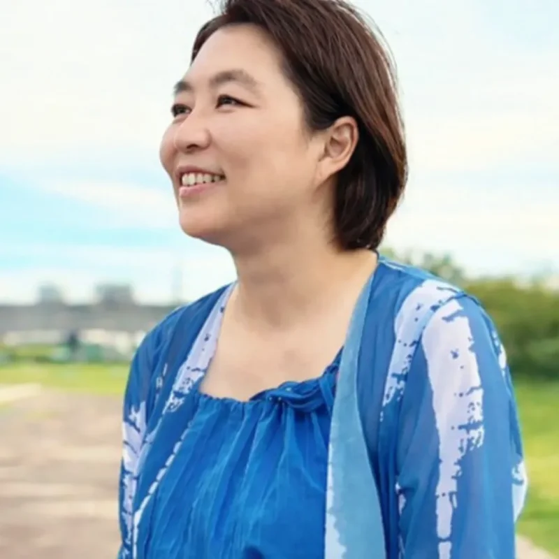 青い服で、川辺の空を見上げて微笑むPLUM